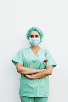 A healthcare professional standing with arms crossed in green scrubs and a protective facemask.